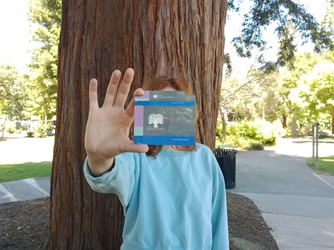 A person holds up a Menlo Park Library card in front of a tree on Burgess campus