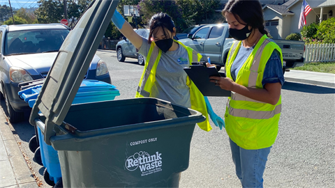 Two individuals in yellow neon vests checking garbage bins