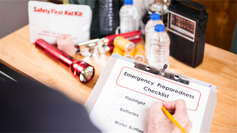 Clipboard that reads Emergency Preparedness Checklist with safety supplies in the background such as water and flashlights