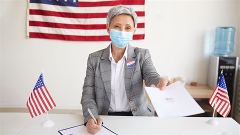 older-woman-wearing-vote-sticker-in-front-of-american-flag-on-the-wall.jpg