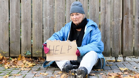 unhoused-older-woman-sits-against-a-fence-holding-a-cardboard-sign-reading-help.jpg